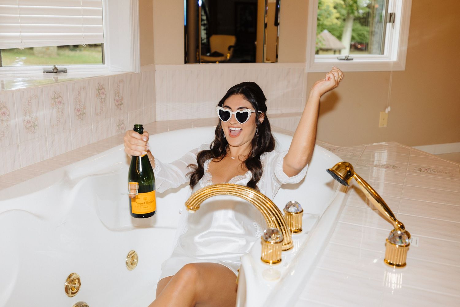 Person relaxing in luxurious bathtub with champagne bottle and gold fixtures wearing sunglasses.