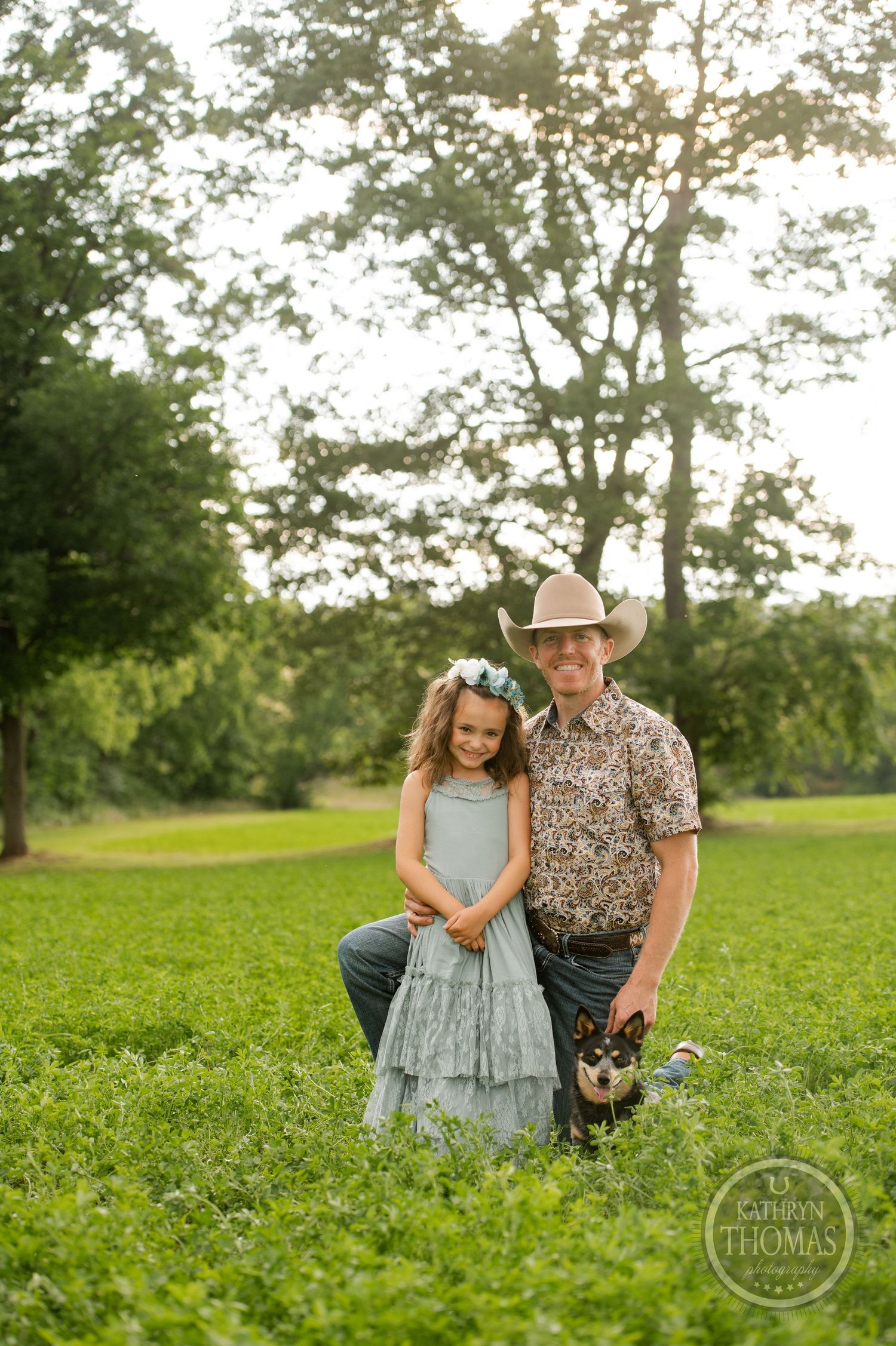 A tender outdoor portrait in a green field with sunlit trees featuring a floral dress and cowboy hat.