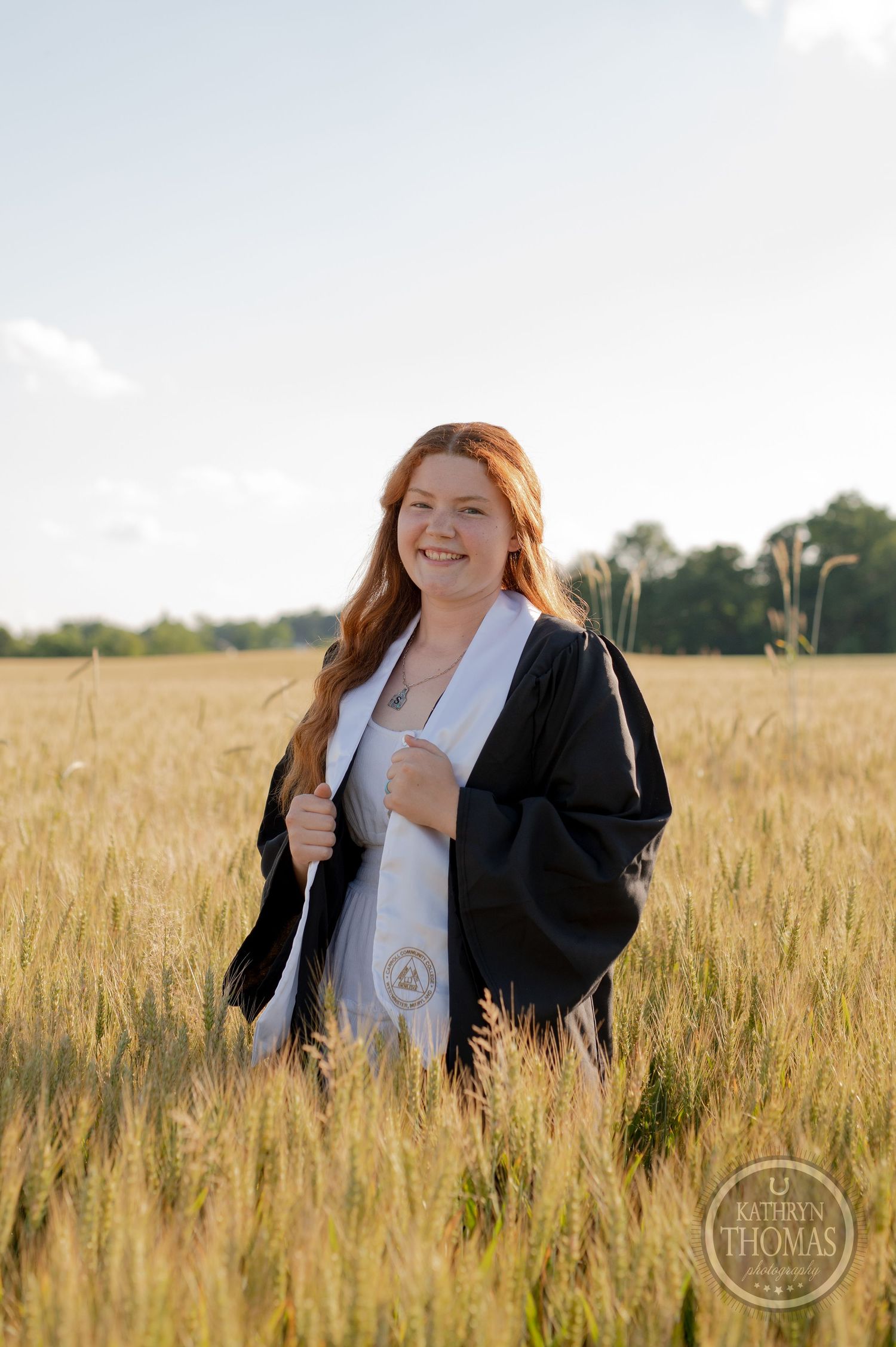 Graduate in academic regalia stands in sun-lit wheat field.