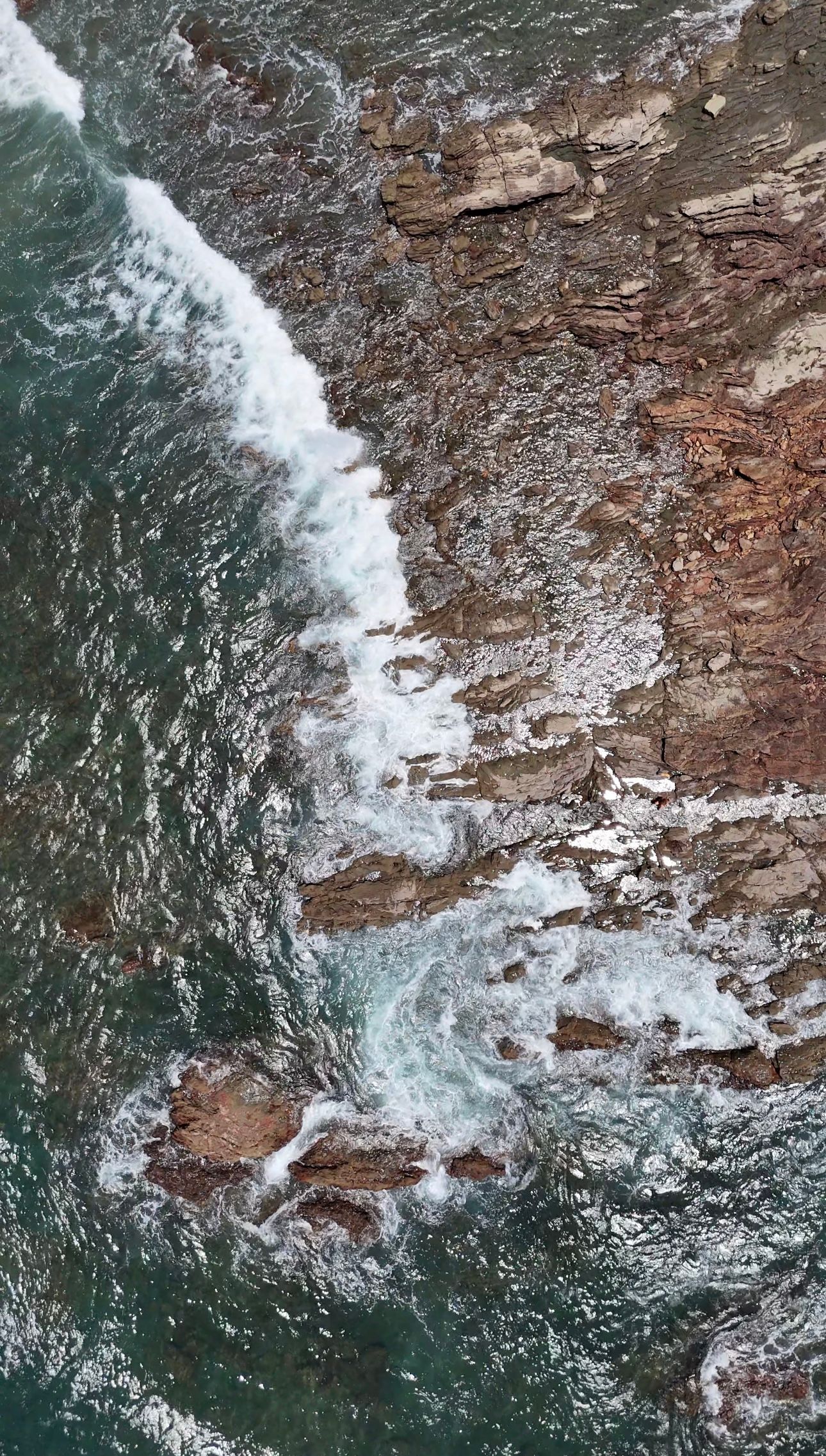 Aerial view of ocean waves crashing against rocky coastal cliffs with turbulent waters.