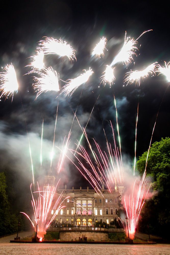 Spectacular fireworks display illuminates historic mansion with red and white pyrotechnics against night sky.