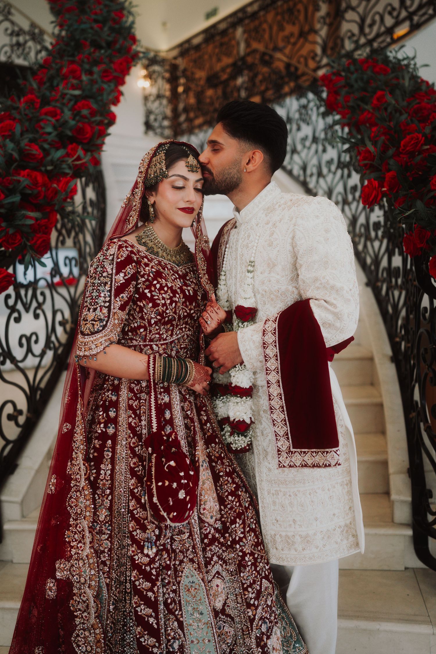 Wedding couple in red lehenga and white sherwani posing by ornate staircase with red roses.