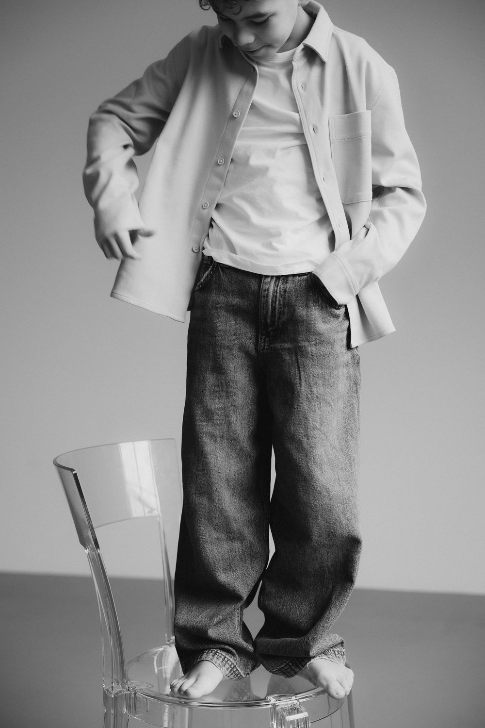 Black and white fashion photo showing someone in casual wear standing on a clear chair wearing jeans and a light jacket.