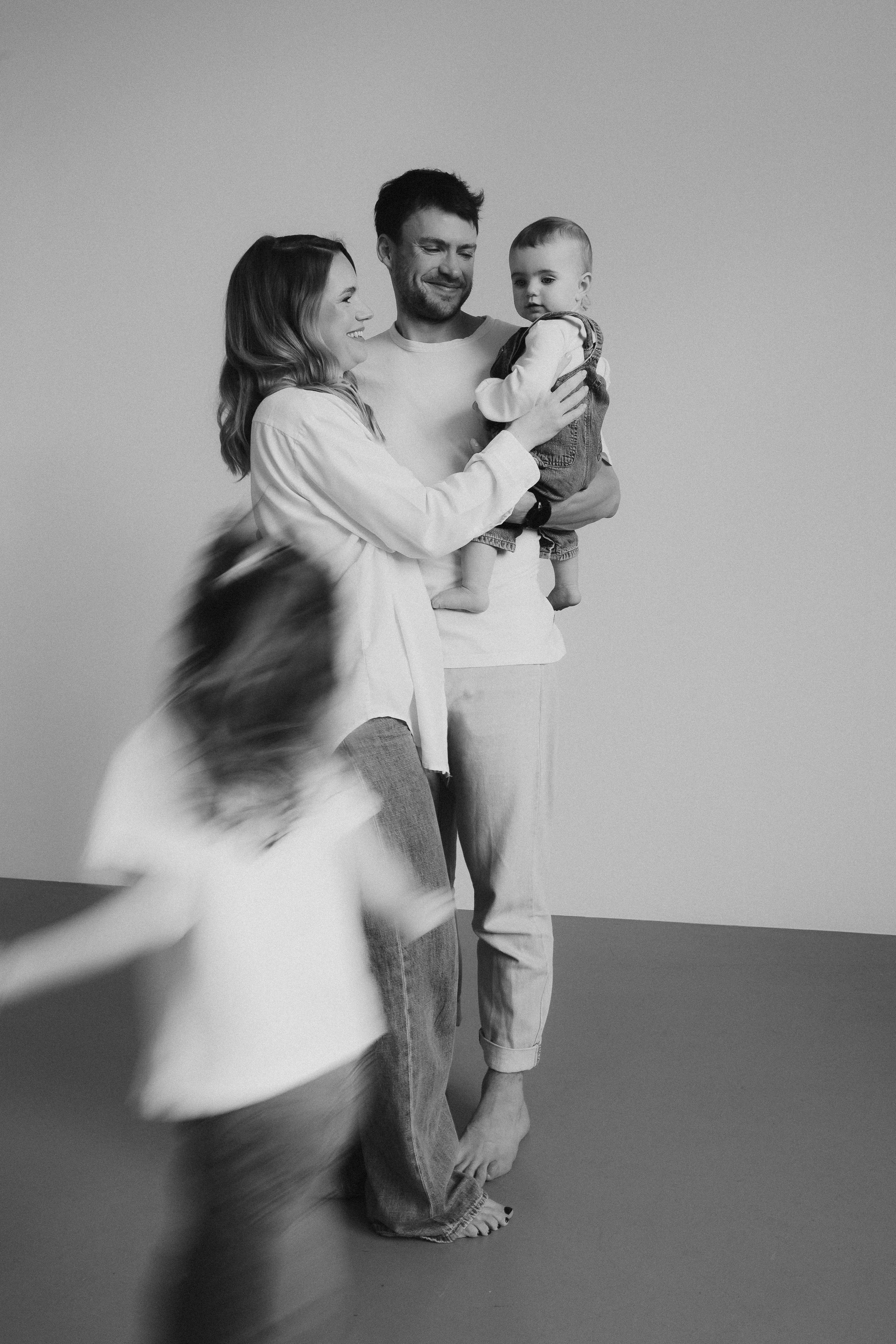 Black and white family portrait with a motion-blurred child running past while parents hold a baby.
