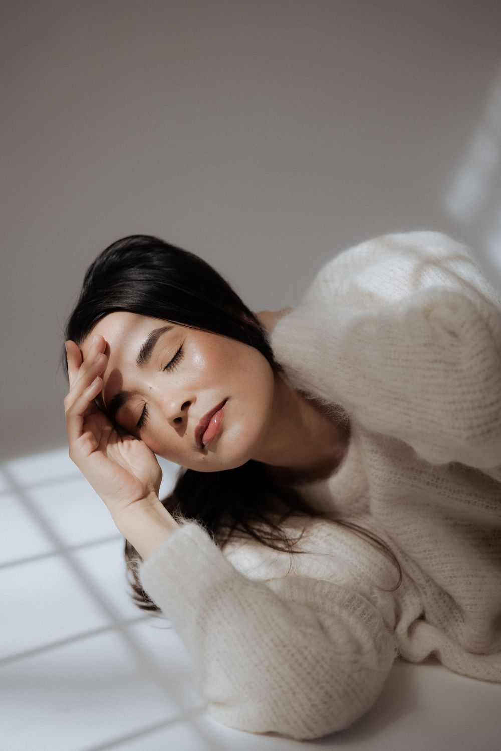 Figure reclining in cream knit sweater with serene expression against minimalist backdrop.