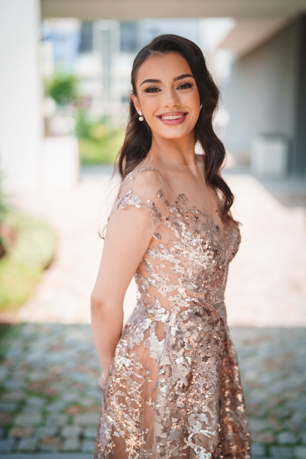 Woman wearing an elegant rose gold sequined evening gown with sheer details and wavy hair smiles outdoors.