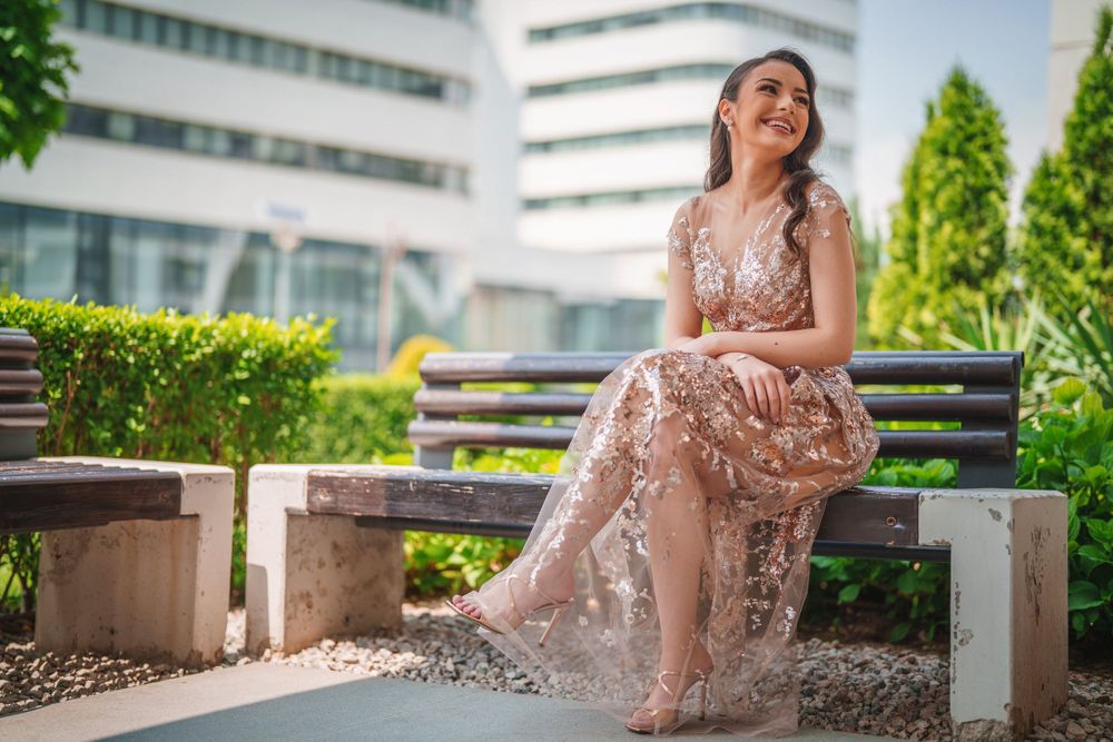 Model in sparkly evening gown poses gracefully on modern concrete bench surrounded by greenery.