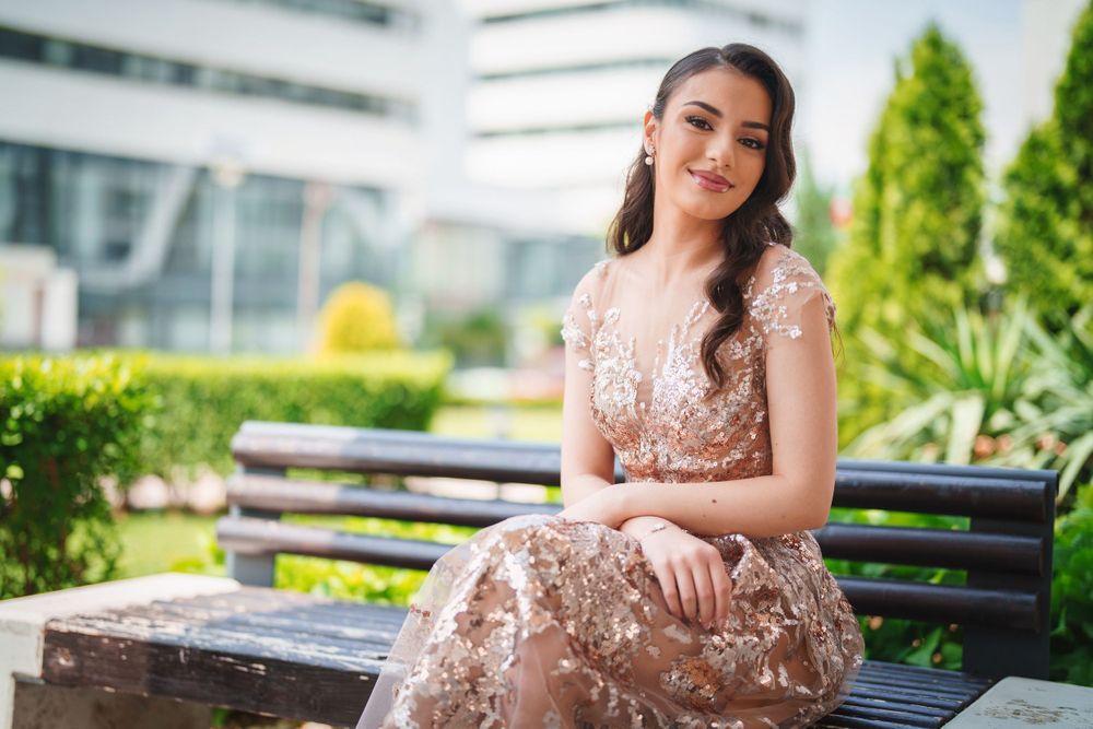 Elegant portrait in rose gold sequined gown seated on bench with modern building backdrop.