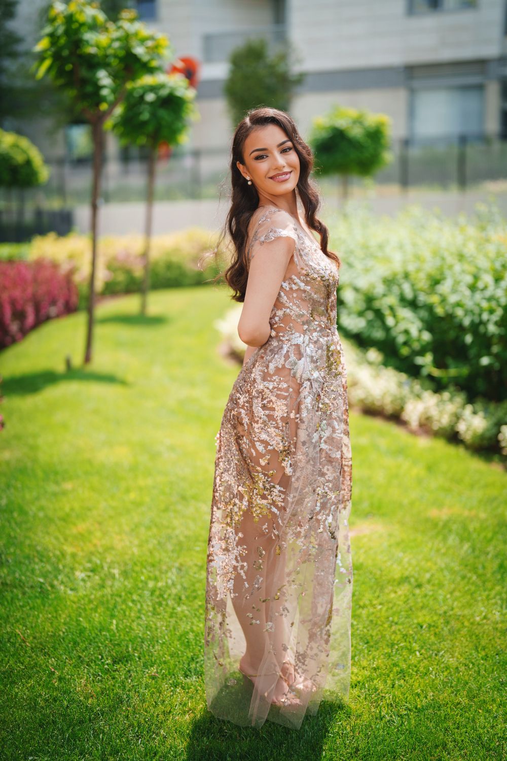 Full-length outdoor portrait in shimmering formal dress amid landscaped garden setting.