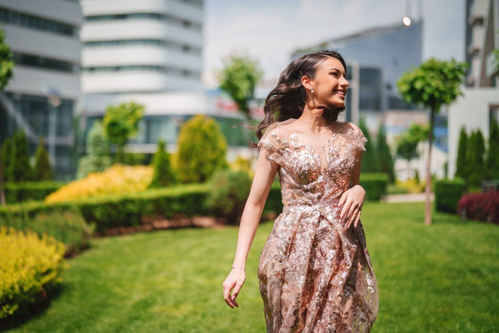 A person in a sparkly rose gold dress twirls joyfully in a garden with modern buildings in the background.