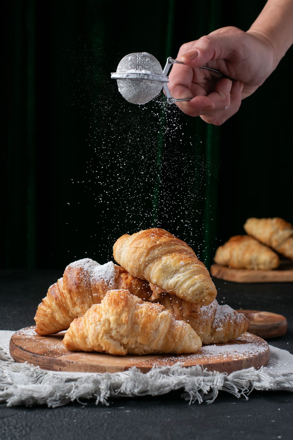 Sprinkling powdered sugar over golden baked croissants on a rustic dark surface.