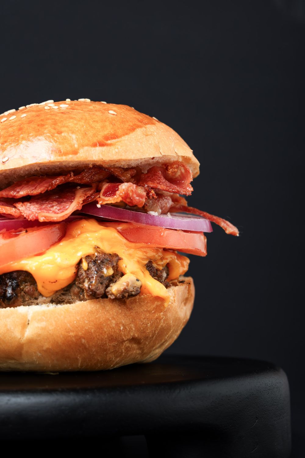 Close-up of juicy cheeseburger with bacon, tomatoes, onions and melted cheese on brioche bun against dark background.