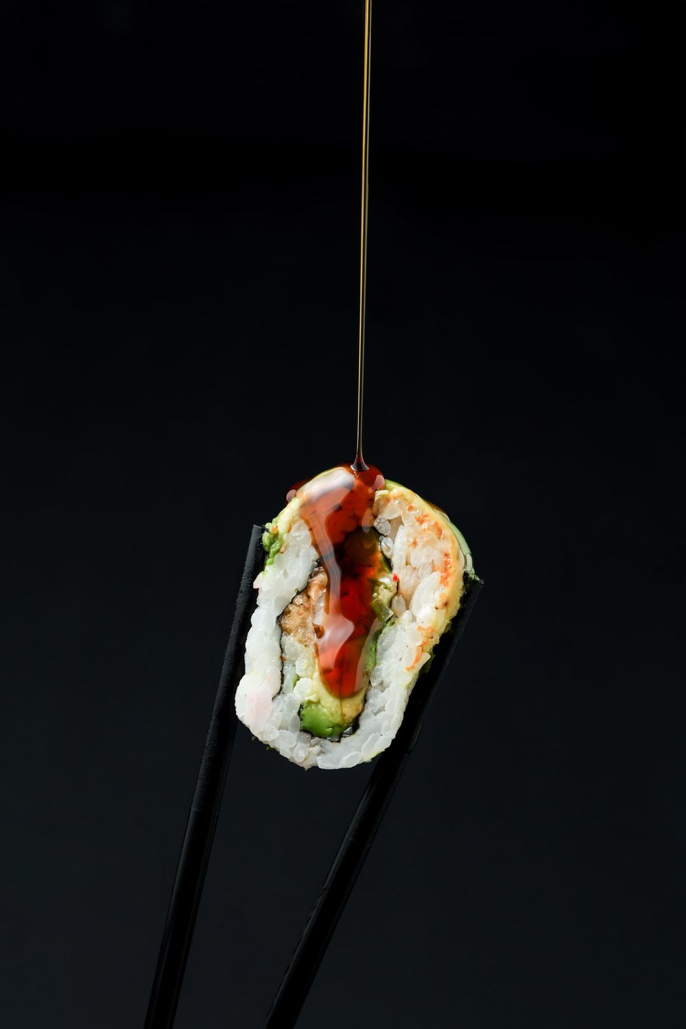 Sushi roll with shrimp and vegetables held by black chopsticks against dark background with soy sauce dripping down.