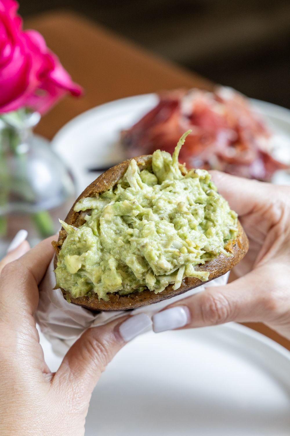Hands holding a slice of toast topped with creamy mashed avocado spread beside pink flowers on a white plate.
