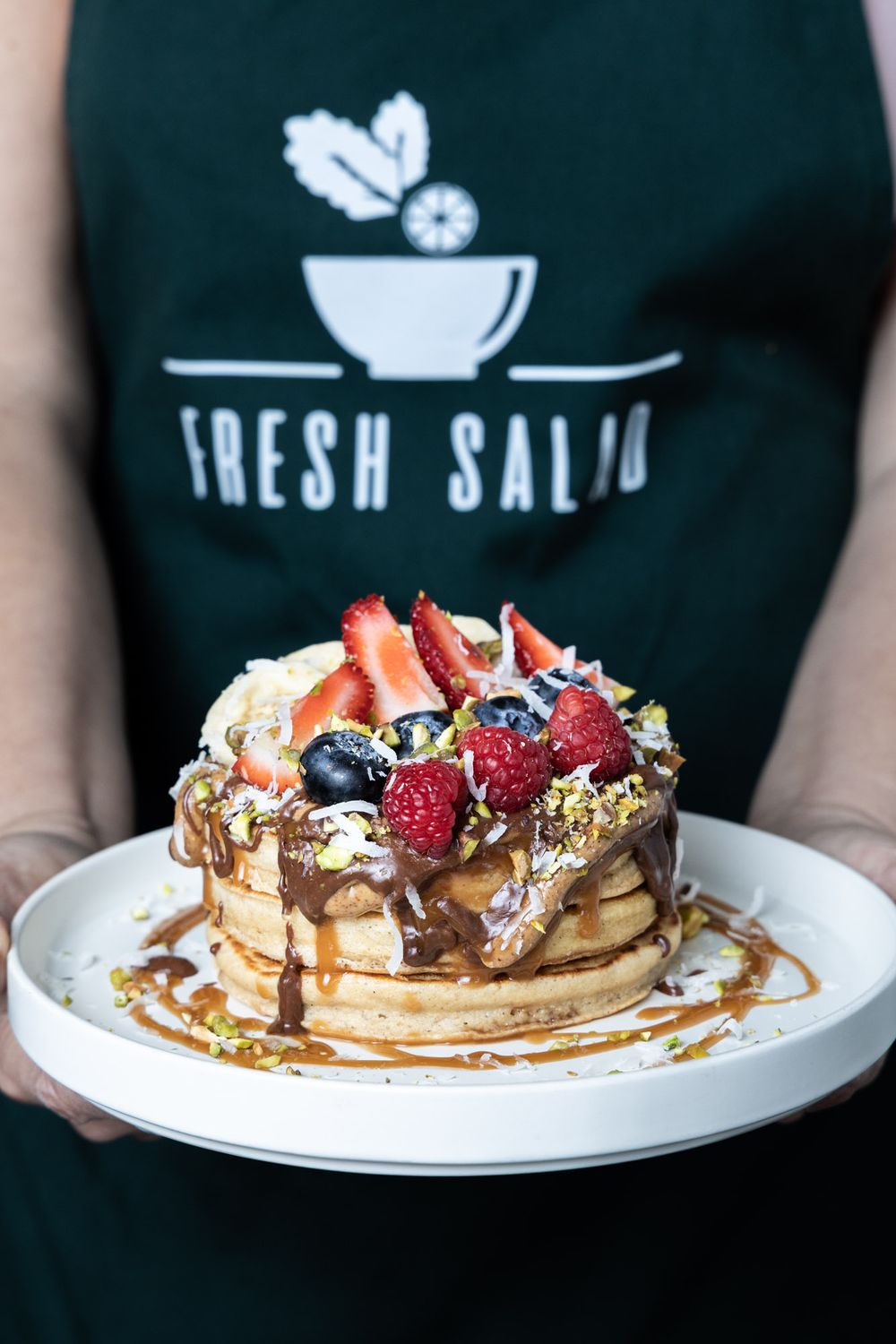 Stack of pancakes topped with fresh berries, whipped cream and chocolate sauce on a white plate.