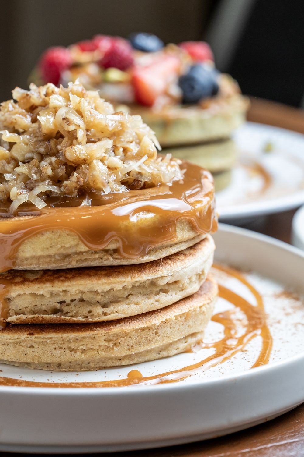 Stack of fluffy pancakes topped with caramel sauce, toasted coconut flakes, with fruit-topped pancakes in background.