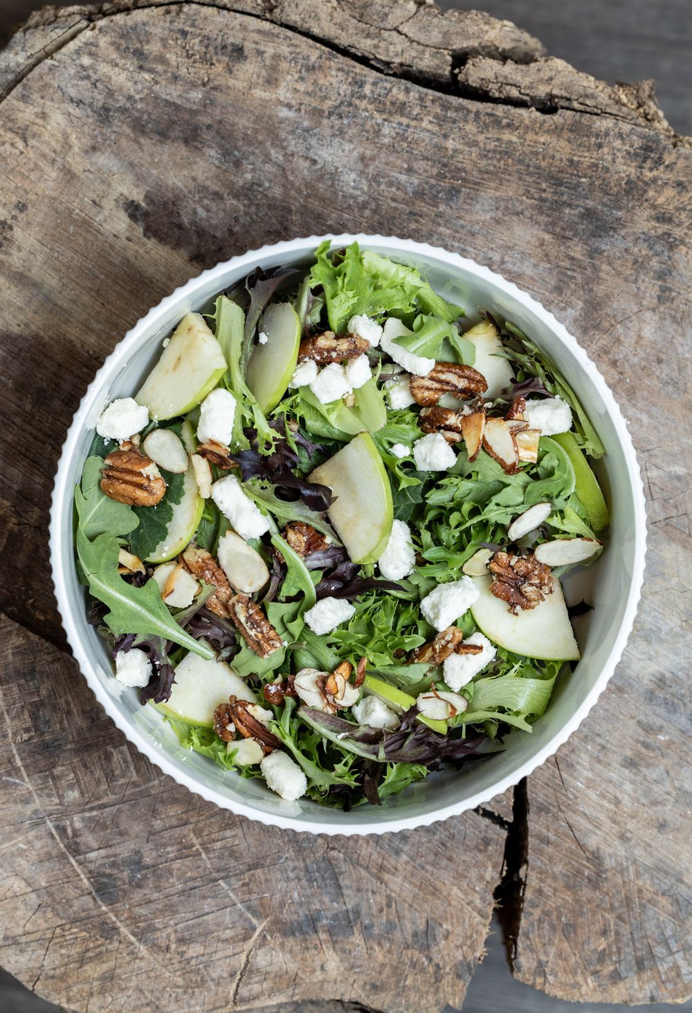 Fresh green salad with crisp apples, pecans, and crumbled goat cheese served in a white bowl on rustic wooden surface.