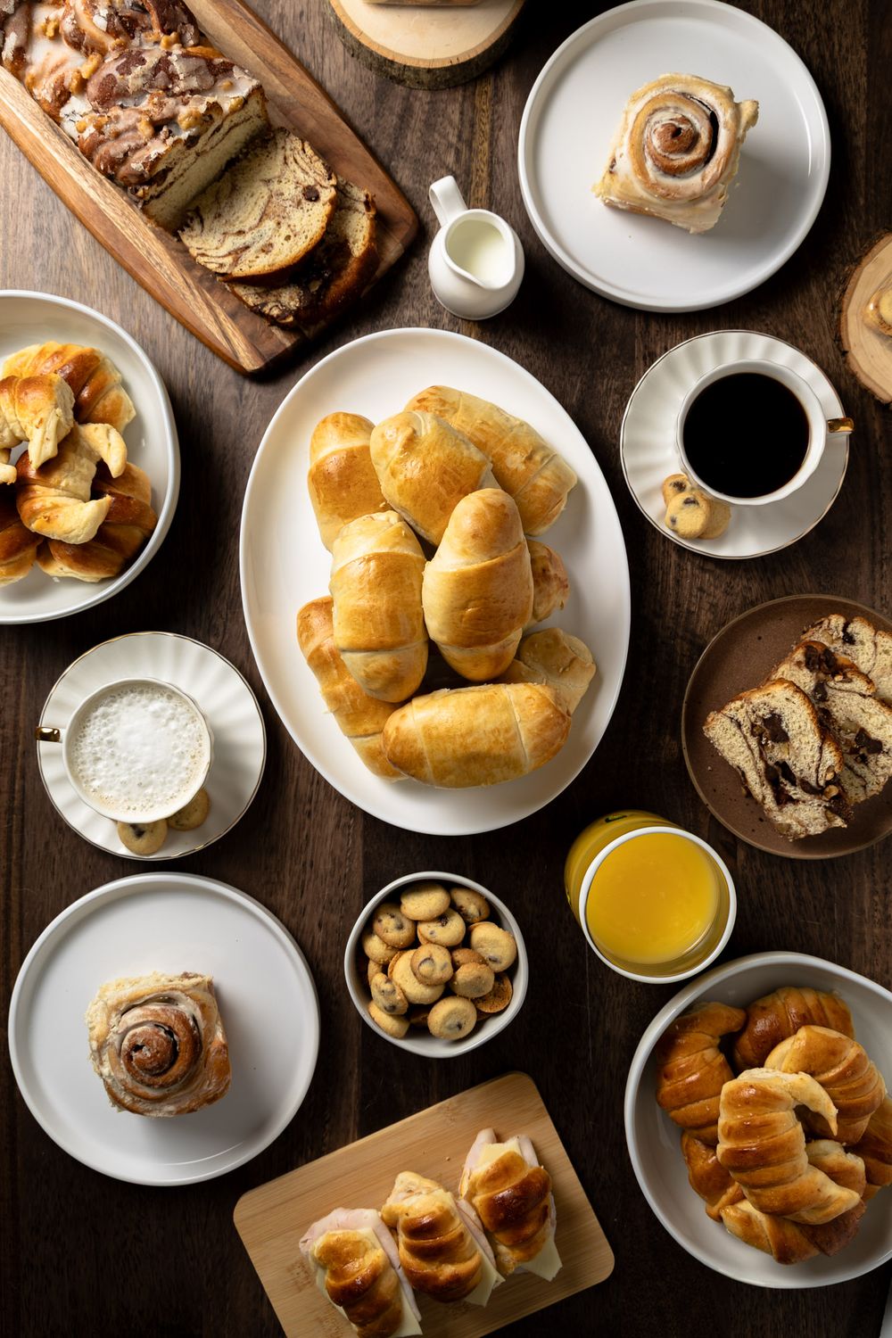 Overhead view of breakfast spread with croissants, cinnamon rolls, coffee, juice and pastries on dark wooden table.