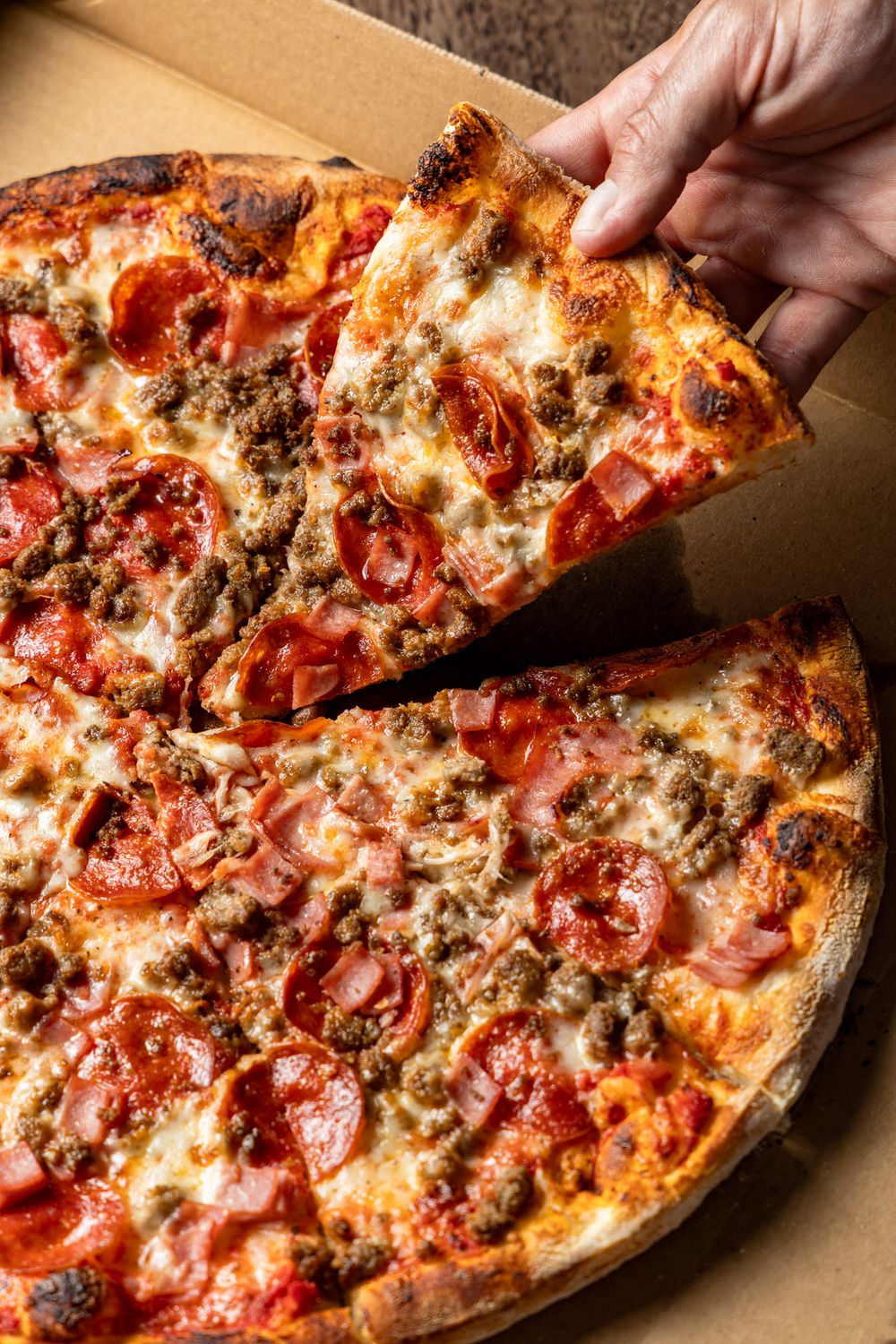 Hot pizza topped with melted cheese, spicy pepperoni and seasoned ground beef being pulled from cardboard box.