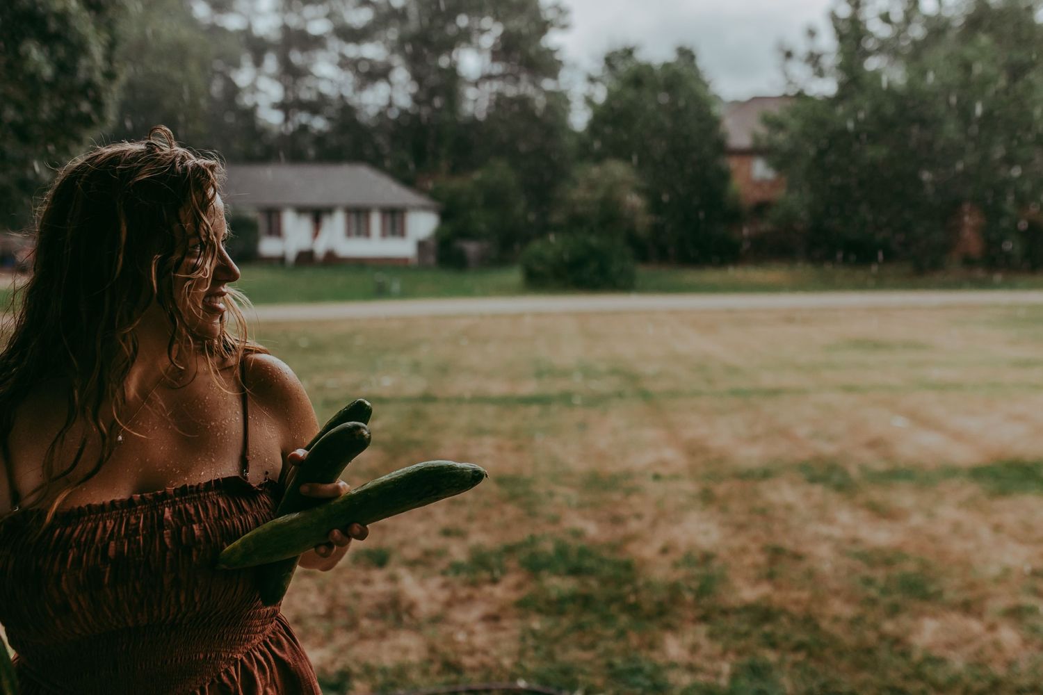 A woman looks out over her yard after running in with cucumber she picked in a summer rain.