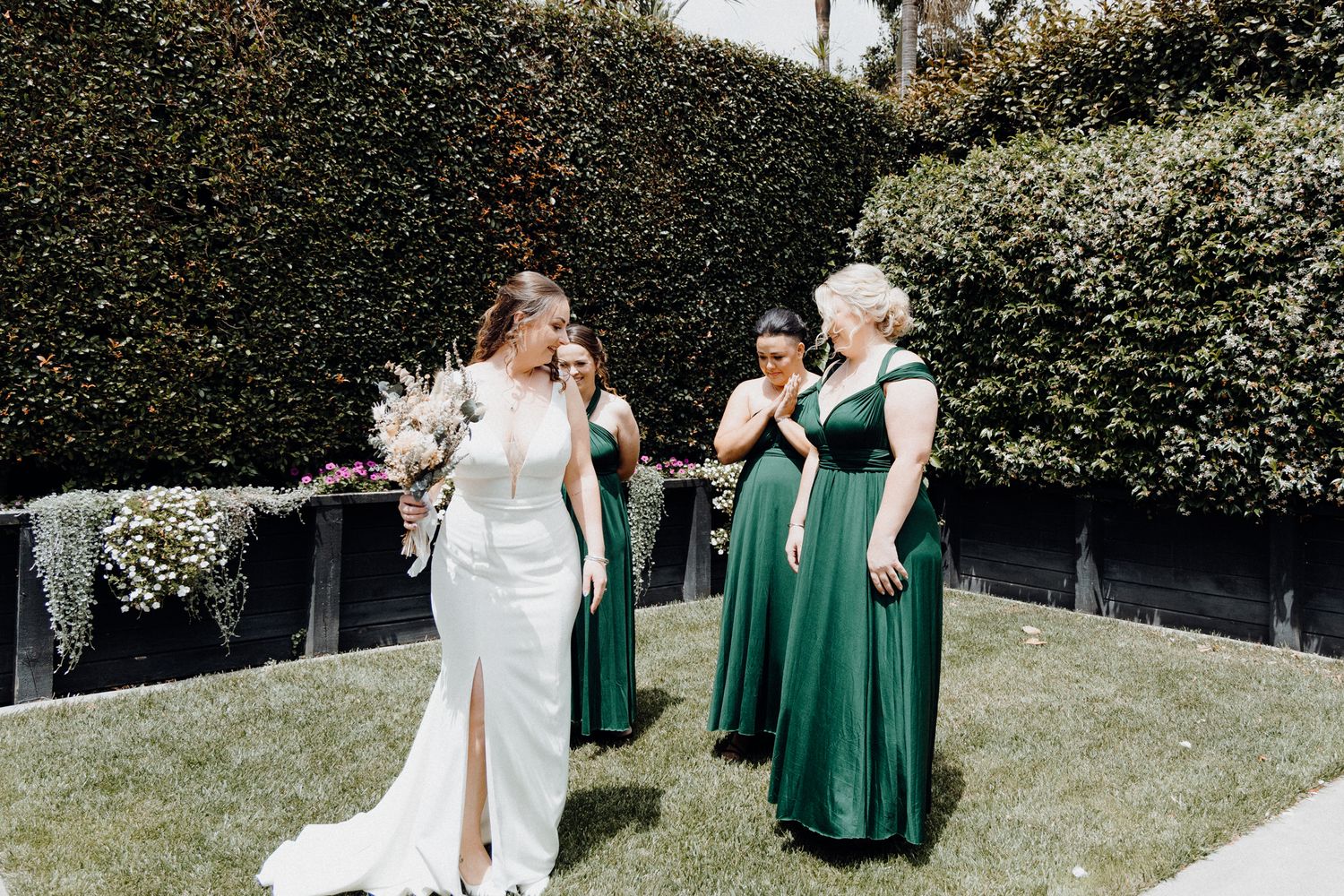 The Bride's Squad laughs together in a sunny garden setting, captured by Apertured Media Auckland Wedding Photographer.