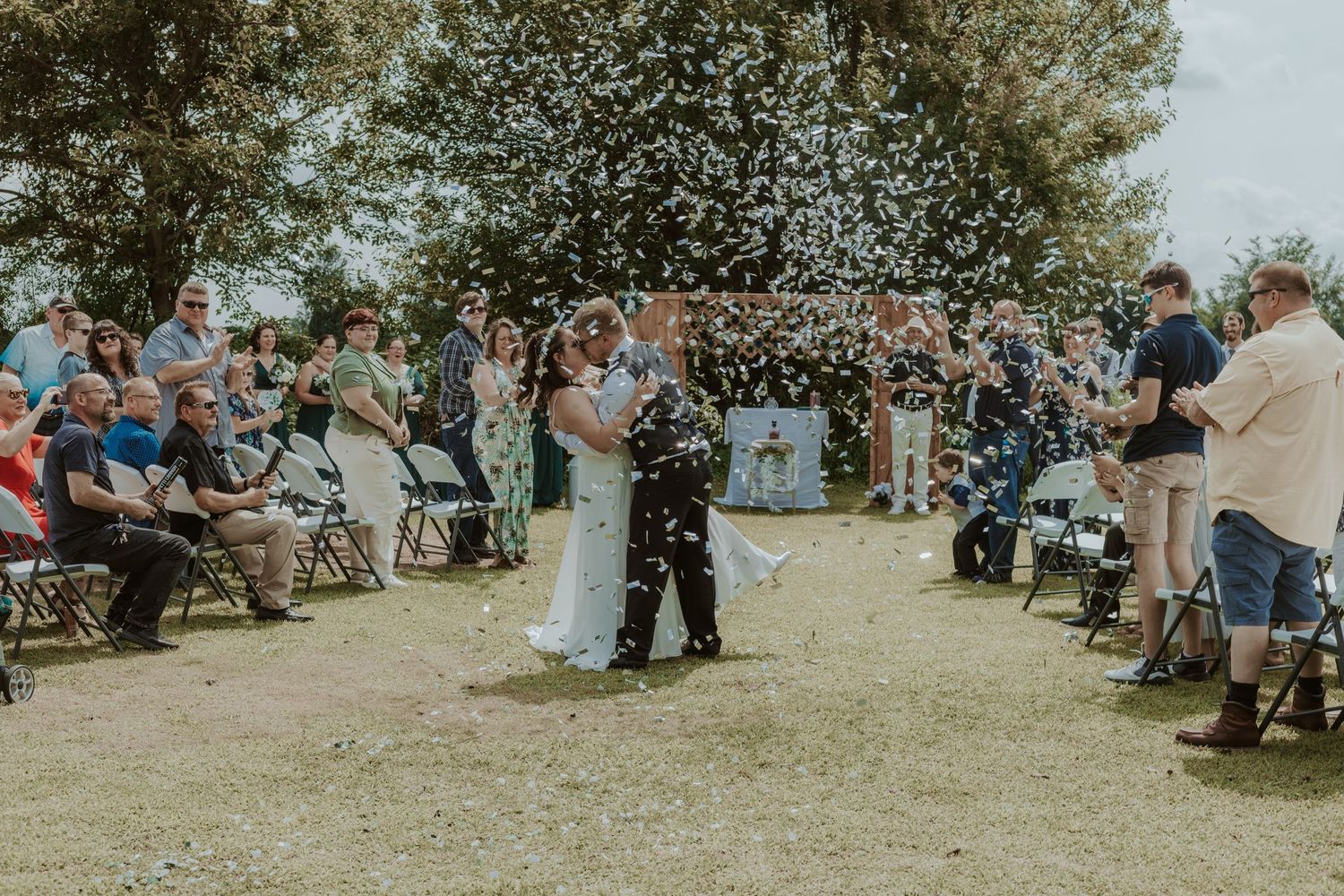 An outdoor wedding ceremony with guests seated on either side of a central aisle under tall trees.
