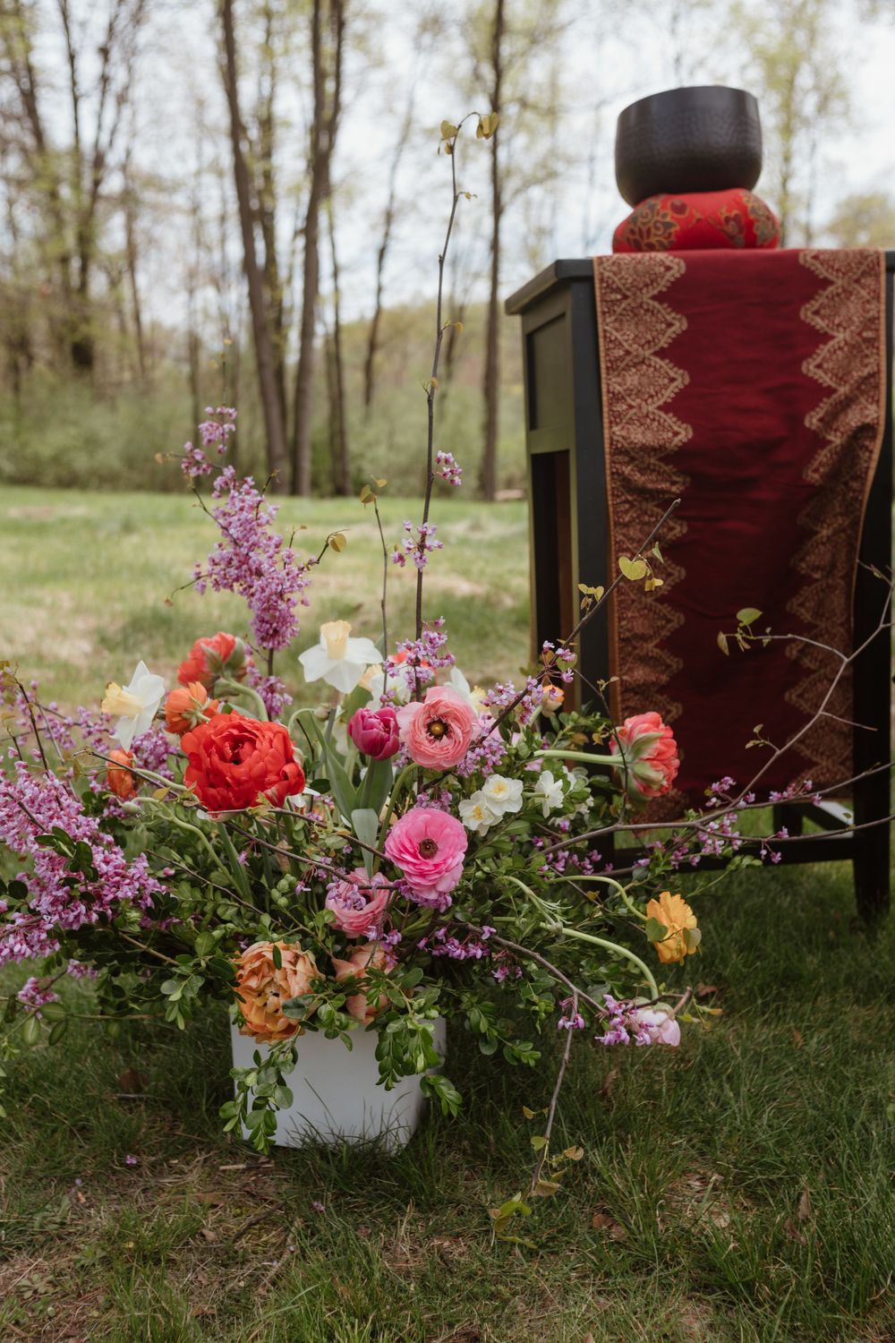 A vibrant flower arrangement with pink and red blooms sits beside an antique style chair with red upholstery outdoors.