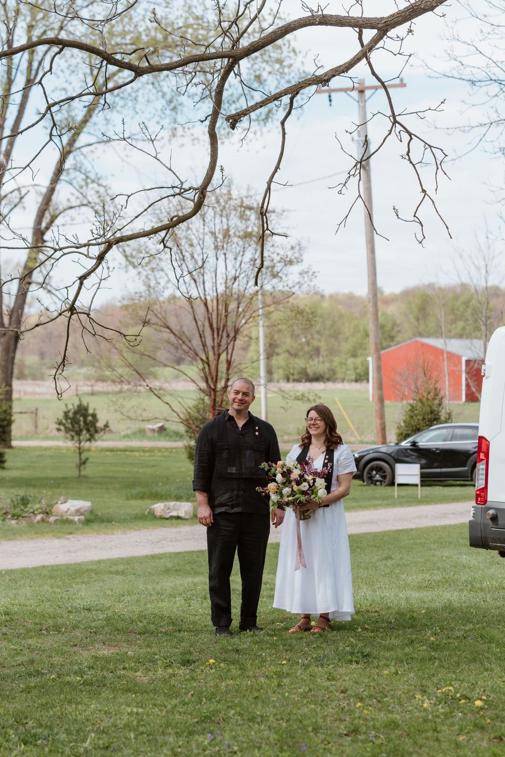 A couple stands together on a grassy lawn with a red barn in the background during a spring day.