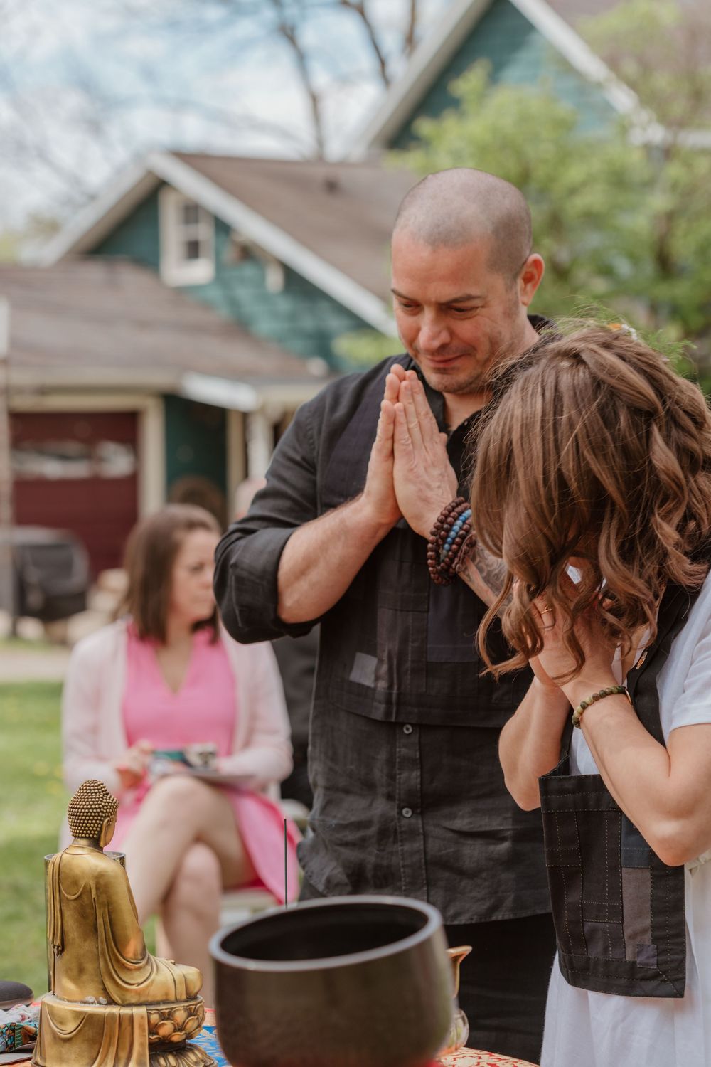 A spiritual ceremony takes place outdoors with a golden buddha statue and meditation bowl in a backyard setting.
