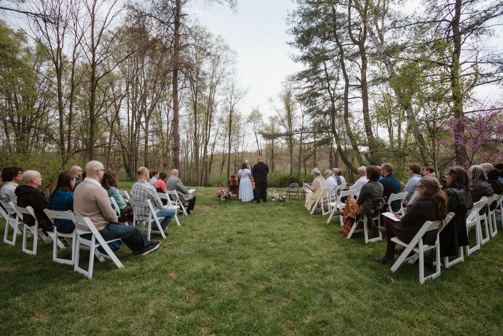 An outdoor wedding ceremony in a wooded setting with white chairs arranged on a grassy lawn for guests.