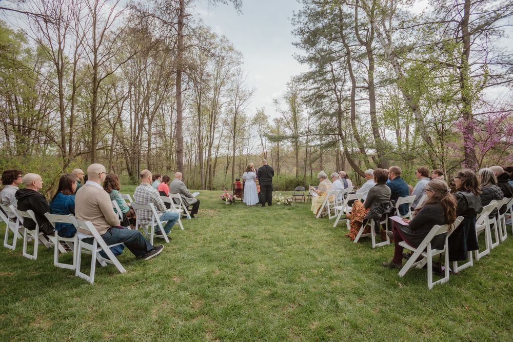 A scenic outdoor wedding ceremony with guests seated on white chairs among spring trees and blossoms.