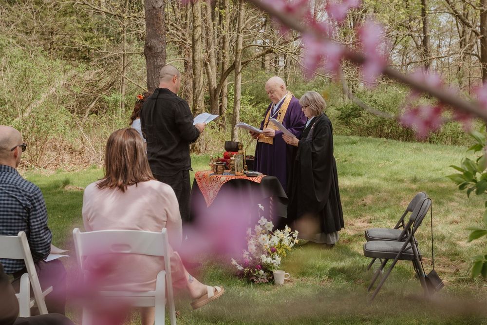 An intimate outdoor spring wedding ceremony with cherry blossoms in bloom and guests seated on folding chairs in a wooded setting.
