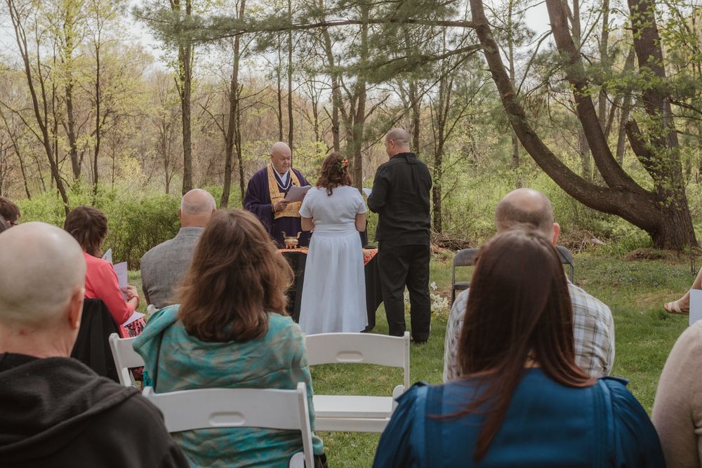 An outdoor wedding ceremony in a wooded setting with guests seated on white chairs watching the proceedings.