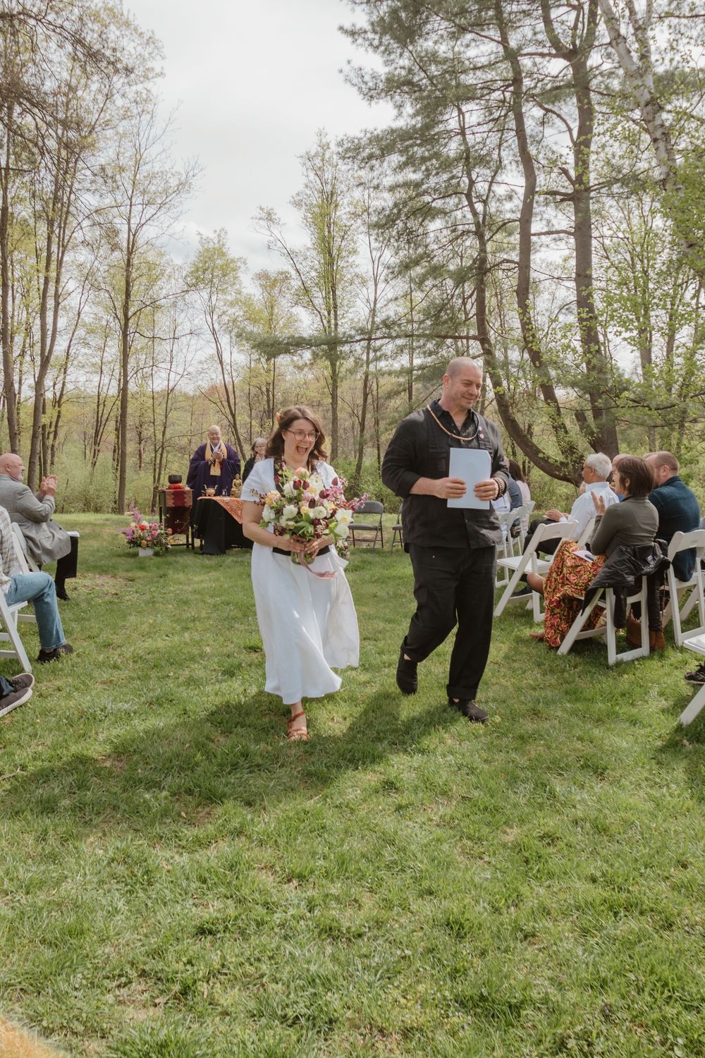 A couple walks down the aisle at their outdoor woodland wedding while guests look on from white chairs.