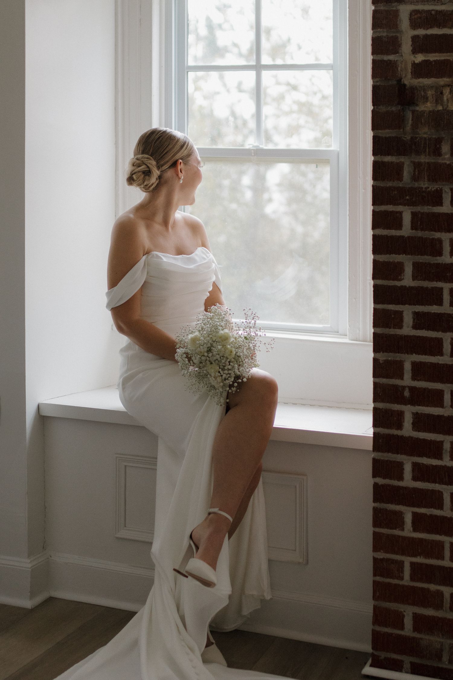 Elegant bride in off-shoulder dress sitting by window with baby's breath bouquet.