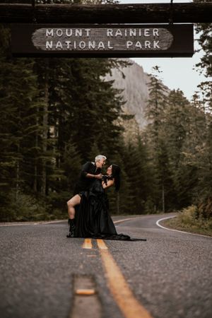 A couple embraces beneath the Mount Rainier National Park entrance sign on a misty road.