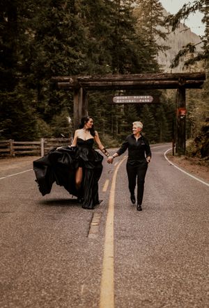 A couple in formal attire walks down a forest road, with the person in a flowing black dress leading the way.