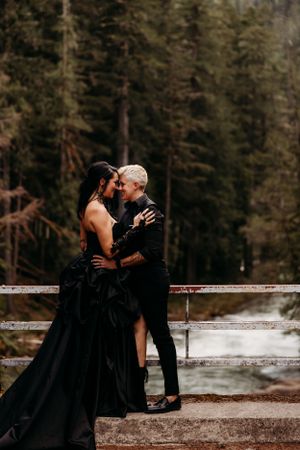 A couple in formal black attire shares an intimate moment on outdoor steps surrounded by forest.