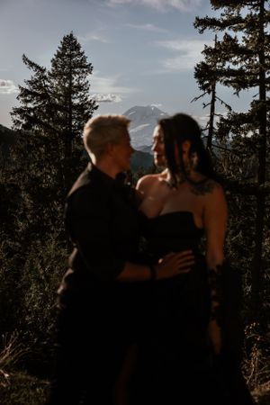 A romantic couple in dark formal wear embraces against a mountain backdrop at sunset.