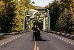 A couple in formal black attire poses on a bridge surrounded by forest scenery.