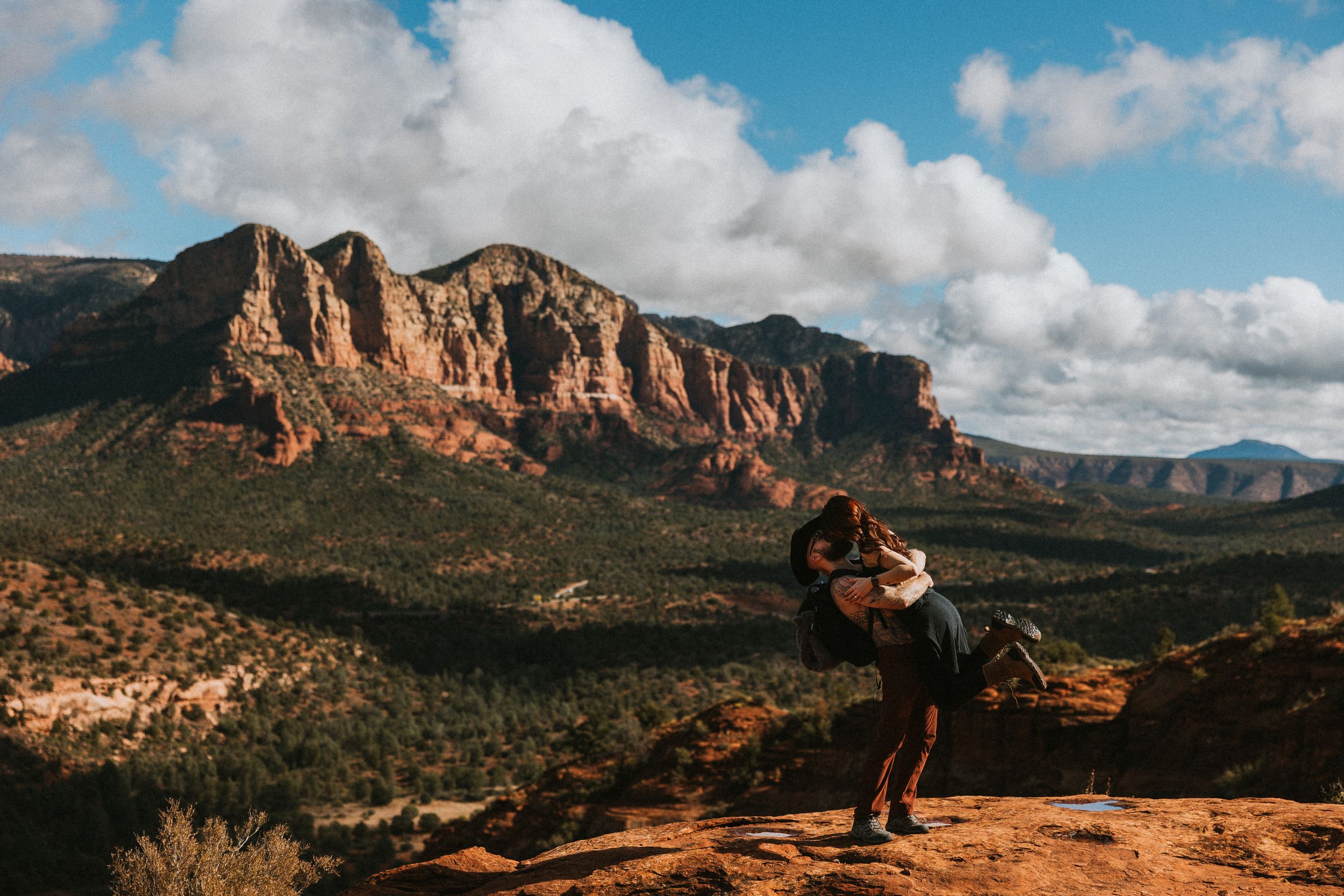 A couple embraces on red rocks with panoramic mountain views in the background.