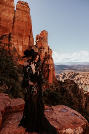 A figure in a black lace dress poses dramatically against red rock formations in a desert landscape.