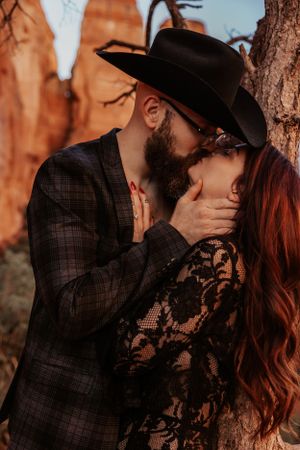 A romantic couple in dark attire share an intimate moment against red rock cliffs at sunset.