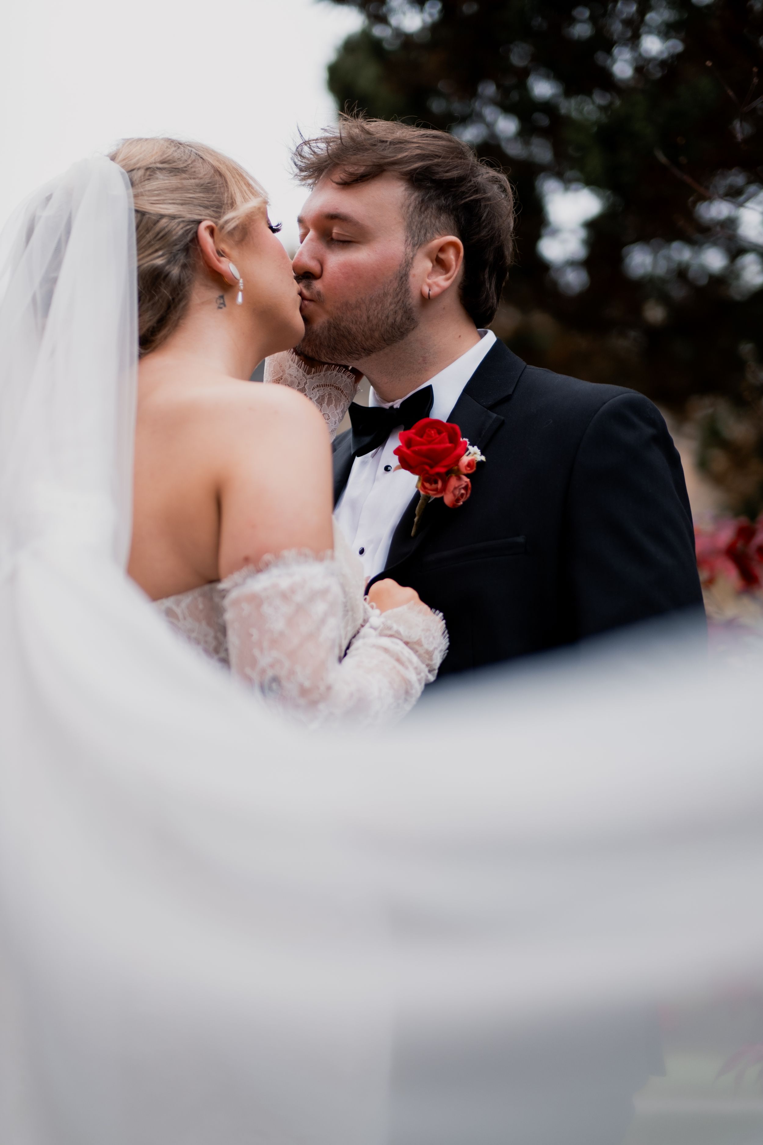 A romantic wedding moment captured through flowing white veil with a red boutonniere visible.