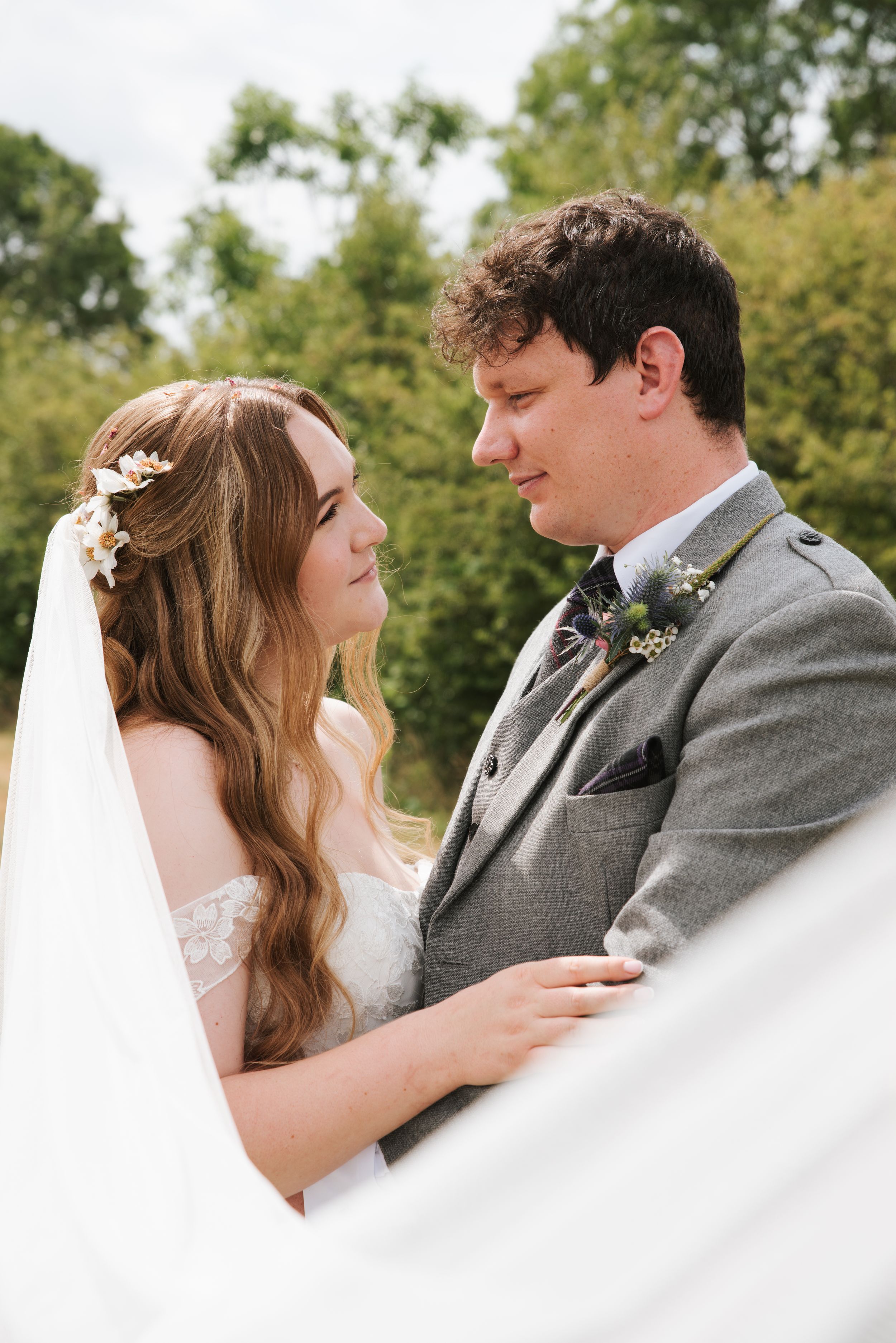 Bride and groom gazing at each other outdoors, she wears floral hair accessories and veil, he wears a grey suit with boutonniere.