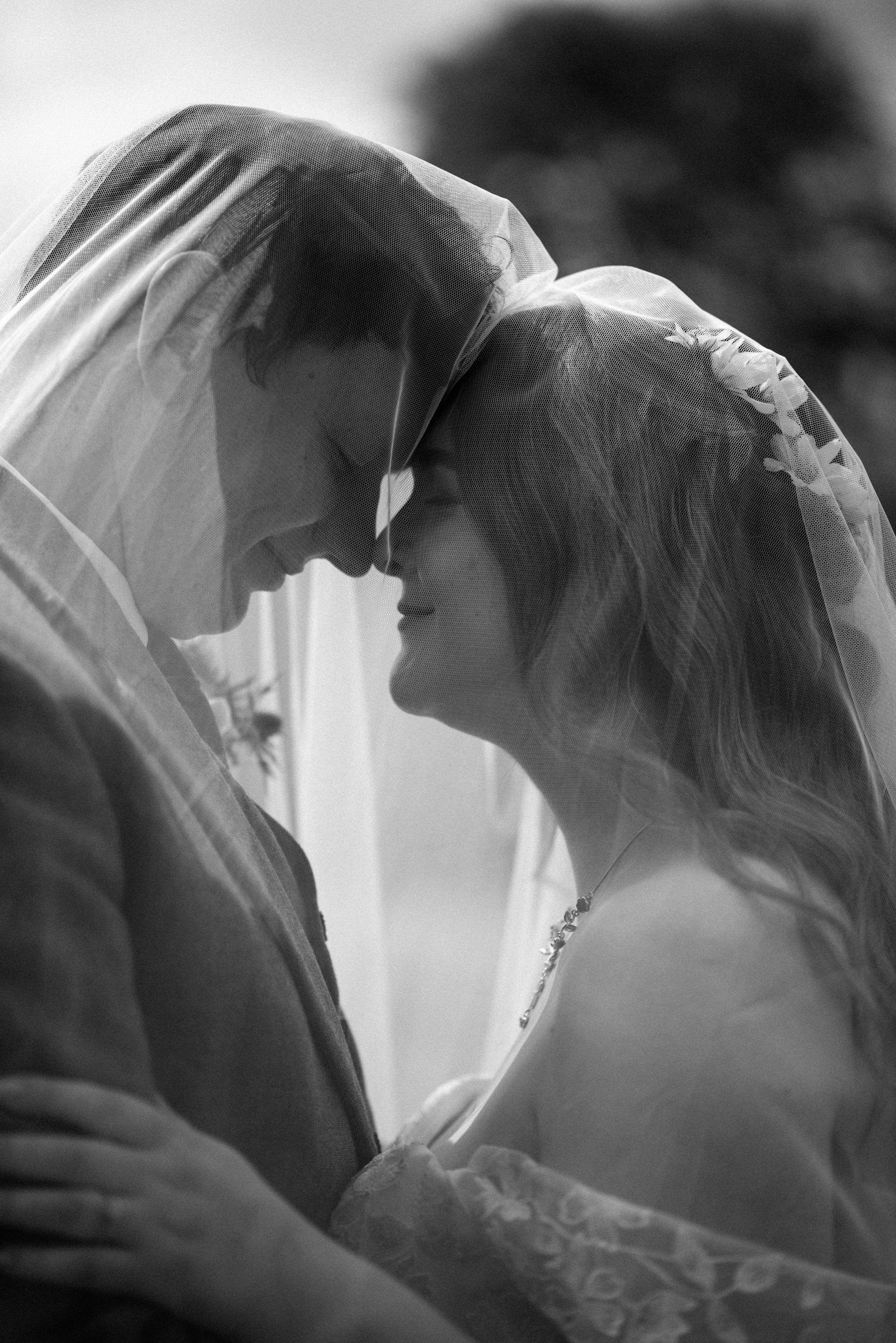 Black and white photo of a bride and groom sharing an intimate moment under a wedding veil.