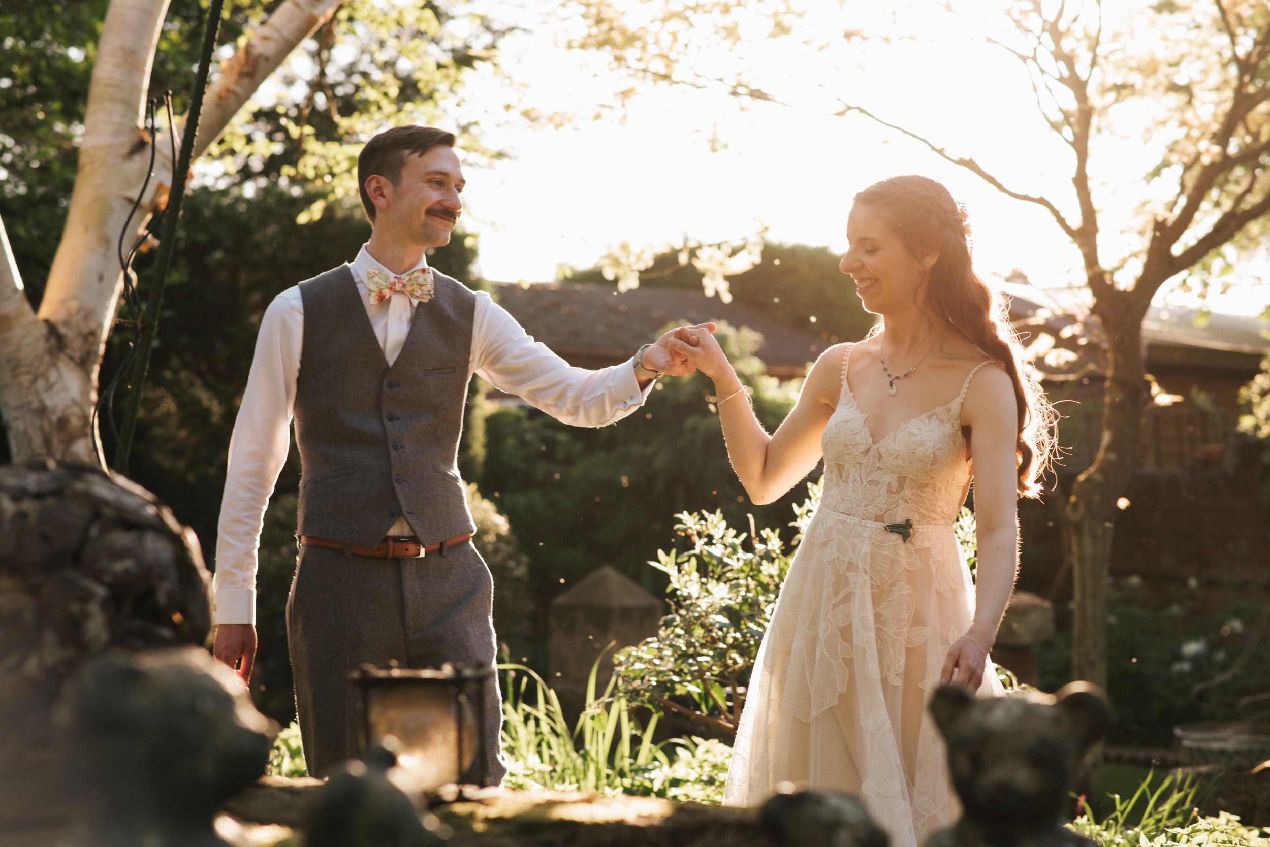Bride and groom dancing outdoors in golden sunlight, wearing elegant wedding attire in a lush garden setting.