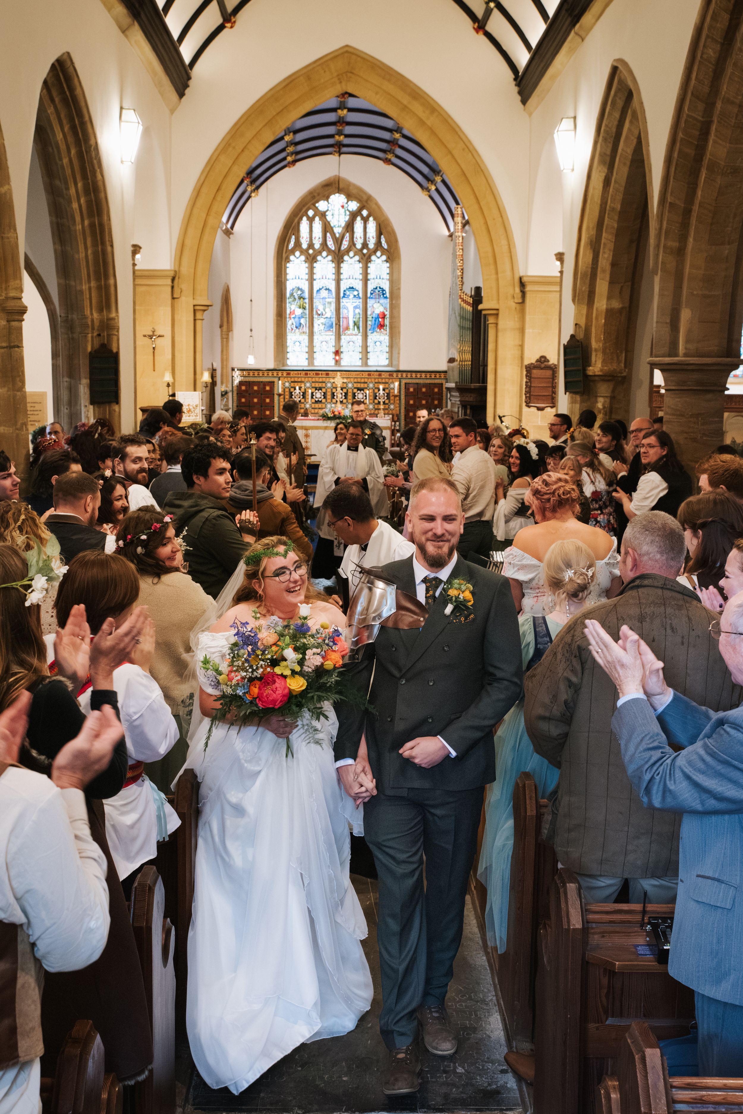 Bride and groom walk down church aisle after wedding ceremony, guests clapping, bride holding colorful bouquet.