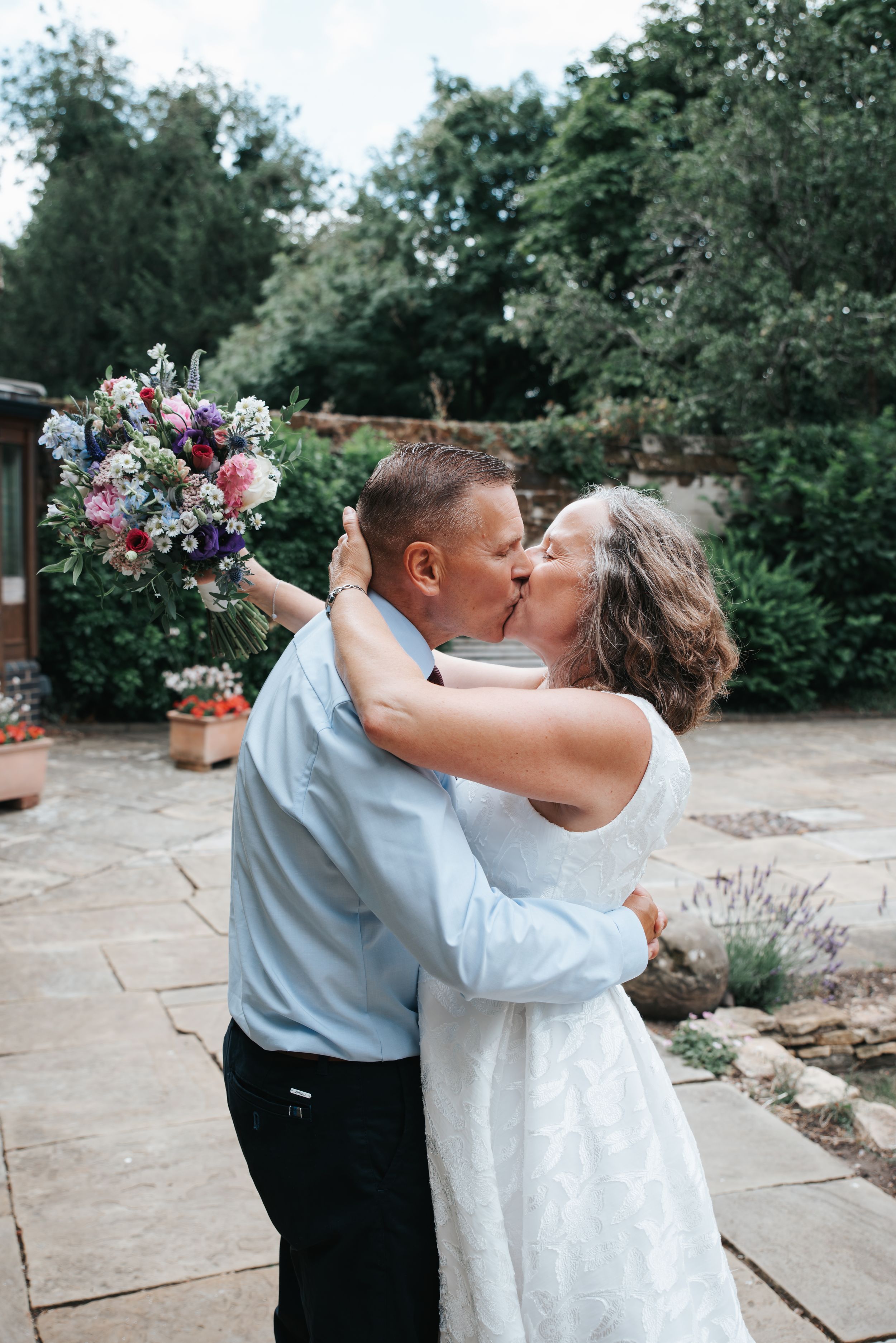 Couple sharing a romantic kiss outdoors on their wedding day, bride in white dress holding colorful bouquet, lush garden background.