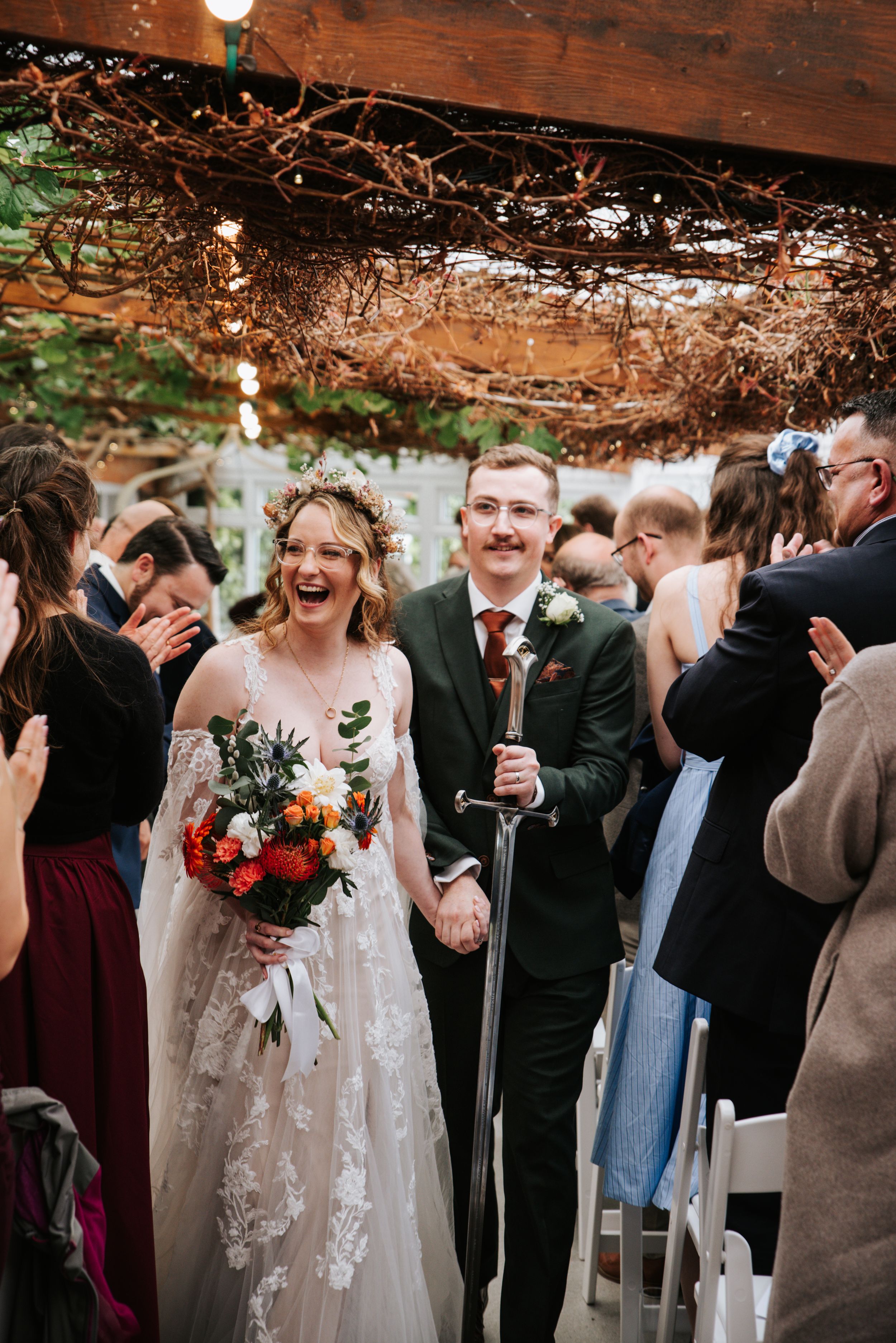 Joyful newlyweds walk down the aisle, bride holding floral bouquet and groom carrying a sword, surrounded by cheering guests.